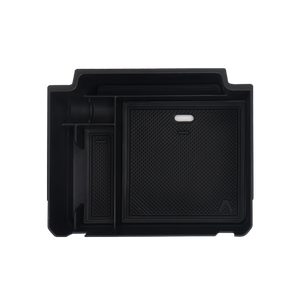 plomor-An image showing the interior of the INEOS Grenadier cab, featuring a custom-fit, black matte ABS storage tray installed on top of the center dashboard. The tray is visible holding small items and shows the built-in cable pass-through slot.