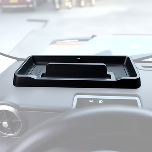An image showing the interior of the INEOS Grenadier cab, featuring a custom-fit, black matte ABS storage tray installed on top of the center dashboard. The tray is visible holding small items and shows the built-in cable pass-through slot.