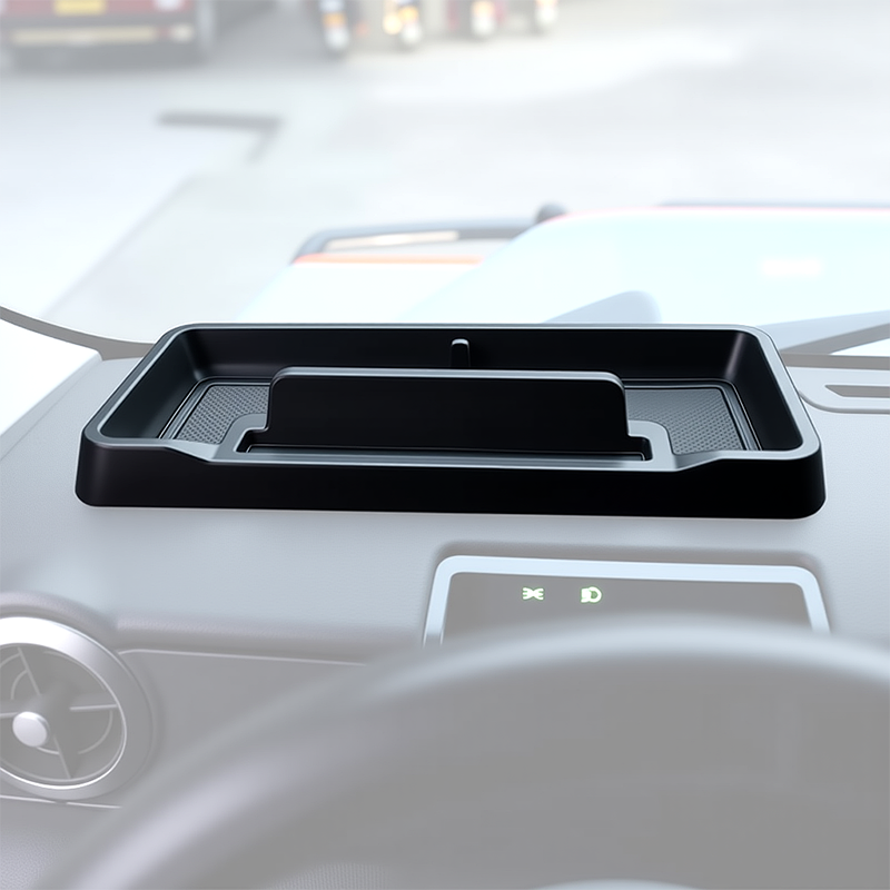 An image showing the interior of the INEOS Grenadier cab, featuring a custom-fit, black matte ABS storage tray installed on top of the center dashboard. The tray is visible holding small items and shows the built-in cable pass-through slot.