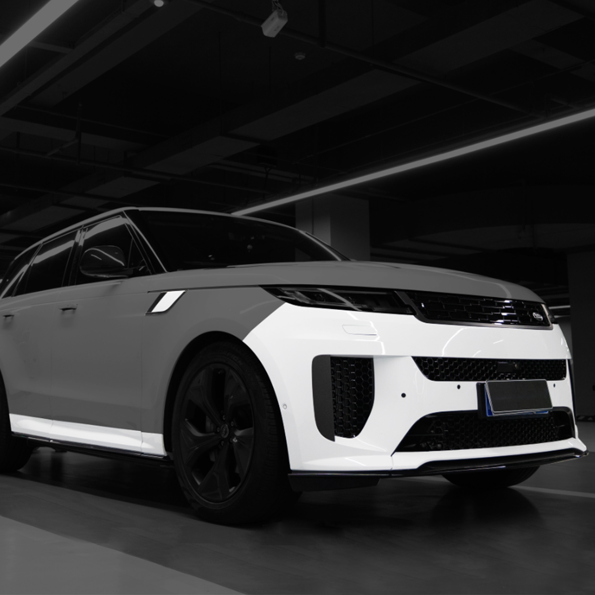 On-vehicle view of PLOMOR front bumper with aero lip and side skirts for Range Rover Sport L461 (2023+), gloss-black accents, installed in indoor garage.